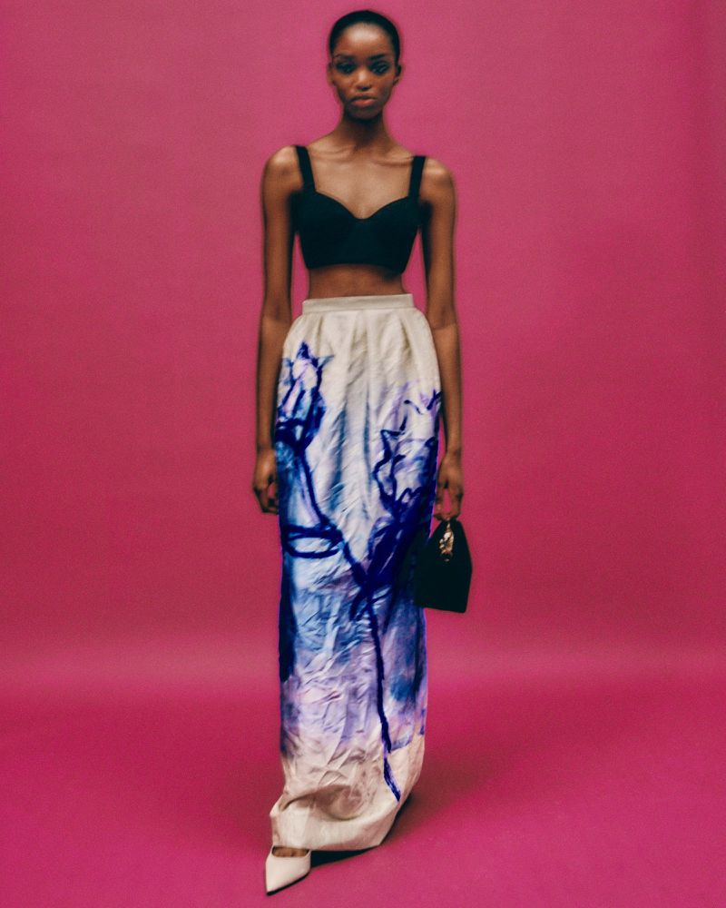 Full-length shot of a Black female model in a black top and long, flowing skirt with a blue design, holding a black purse on a pink backdrop.