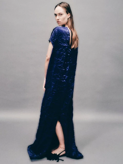 A slender woman with long brown hair poses in a floor-length textured blue dress, glancing back over her shoulder in a studio setting.