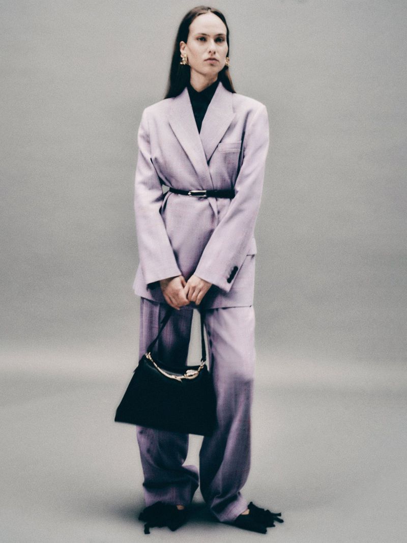 A woman in a light purple suit holds a black bag, posing in front of a grey backdrop in a fashion shot emphasizing modern professional style.