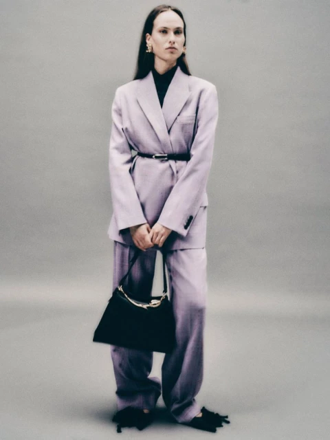 A woman in a light purple suit holds a black bag, posing in front of a grey backdrop in a fashion shot emphasizing modern professional style.