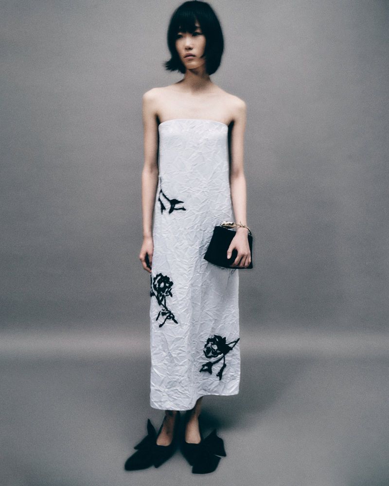 A slender, fair-skinned model in a strapless white midi dress with black rose accents, carrying a black clutch, poses against a gray backdrop.