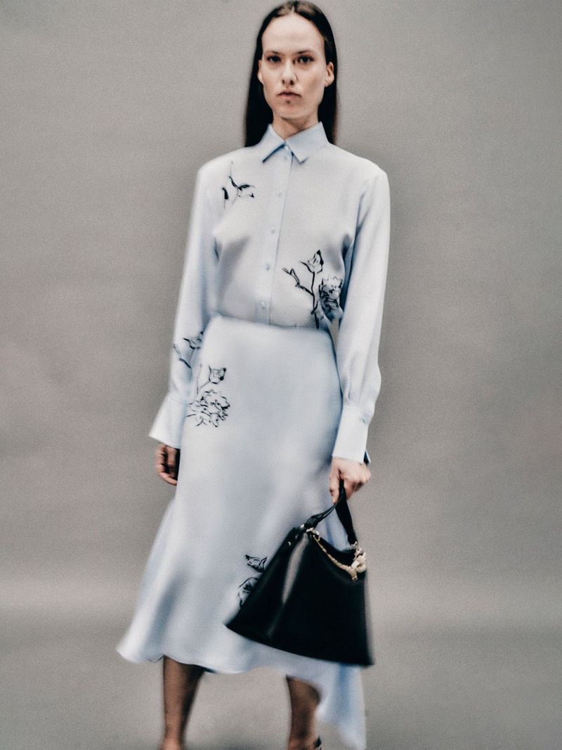 Fashion model in a light blue floral-print dress and black purse poses in a studio setting. Editorial fashion shot.
