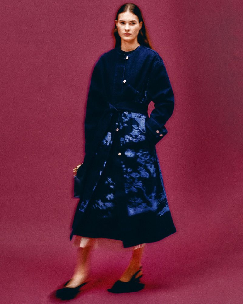 A woman in a blue patterned coat and black slingback heels poses against a magenta background in a fashion studio portrait.