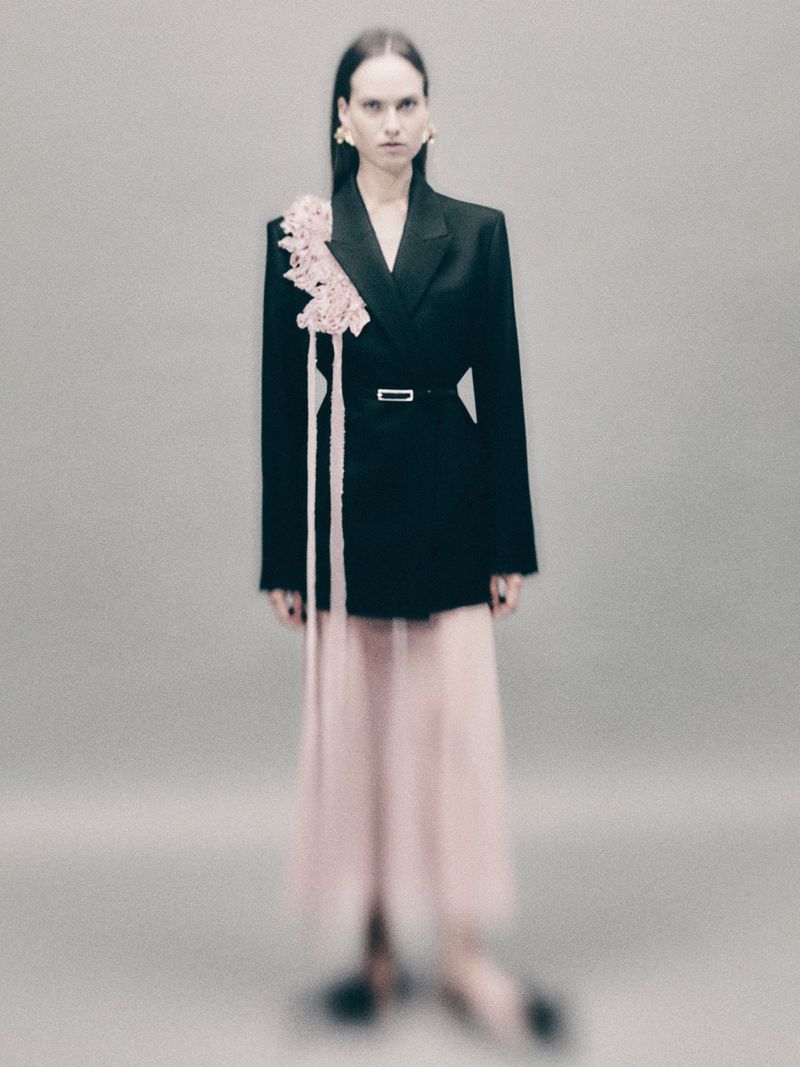 Elegant adult female posing in studio, wearing black blazer with floral detail and flowing pink skirt, sophisticated fashion concept.