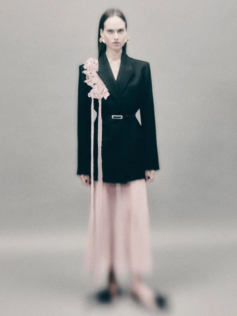 Elegant adult female posing in studio, wearing black blazer with floral detail and flowing pink skirt, sophisticated fashion concept.