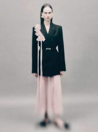 Elegant adult female posing in studio, wearing black blazer with floral detail and flowing pink skirt, sophisticated fashion concept.