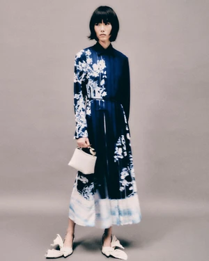 Model in blue and white patterned long dress holding a small handbag against a neutral grey studio background