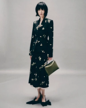 Model with dark bob haircut wearing a black floral jacket and skirt holding a green handbag against a plain grey background