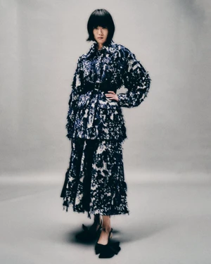 Model with dark bob haircut wearing a blue and black patterned belted jacket and matching skirt against grey seamless studio background.