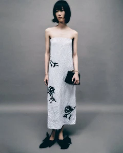 Model wearing a strapless white floral dress holding a black handbag against a grey studio backdrop