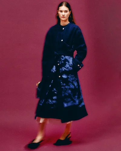 Model in a dark patterned midi coat and black heels against a deep burgundy backdrop for Erdem Pre-Fall 2025 lookbook.