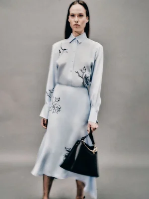 Model wearing a light blue matching shirt and skirt set with floral embroidery, holding a black handbag against a grey backdrop