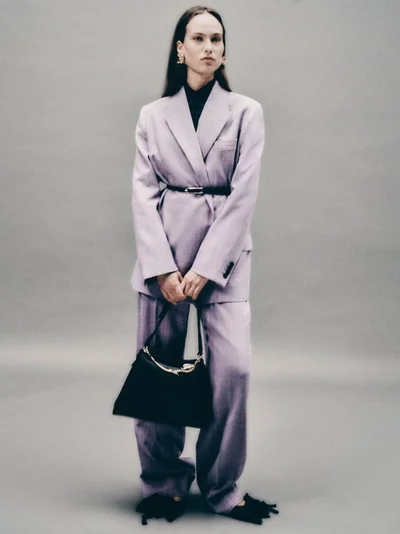 Model wearing a lavender pantsuit and black handbag against a grey seamless studio backdrop.