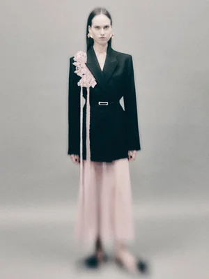 Model wearing a black blazer with pink floral appliqués over a sheer pink skirt against a grey background.