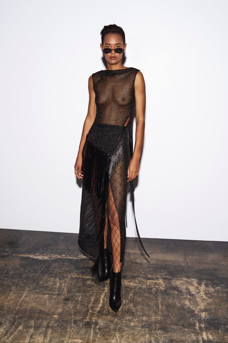 Fashionable woman poses in studio wearing a black net dress, sunglasses, and ankle boots against a stark white background.