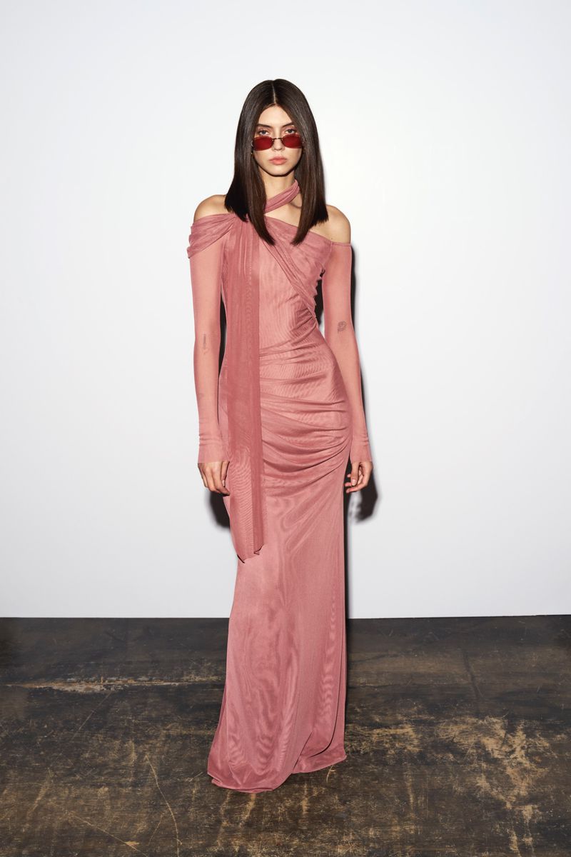 Model wearing a dusty rose halter neck long dress and red sunglasses standing against a plain white studio wall