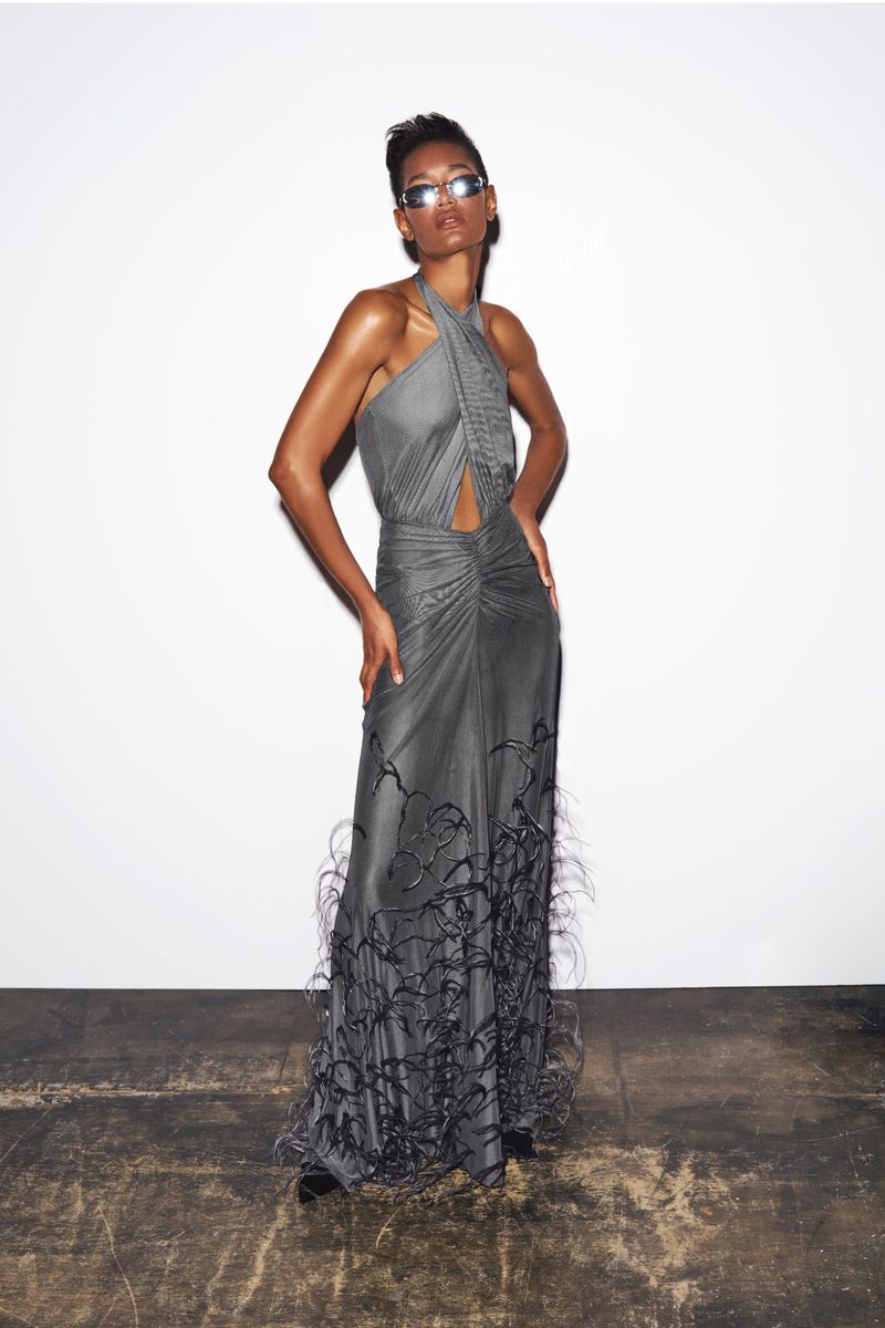 An adult woman poses in a sleeveless, grey gown with feather accents against a white wall and dark floor, showcasing fashion style.