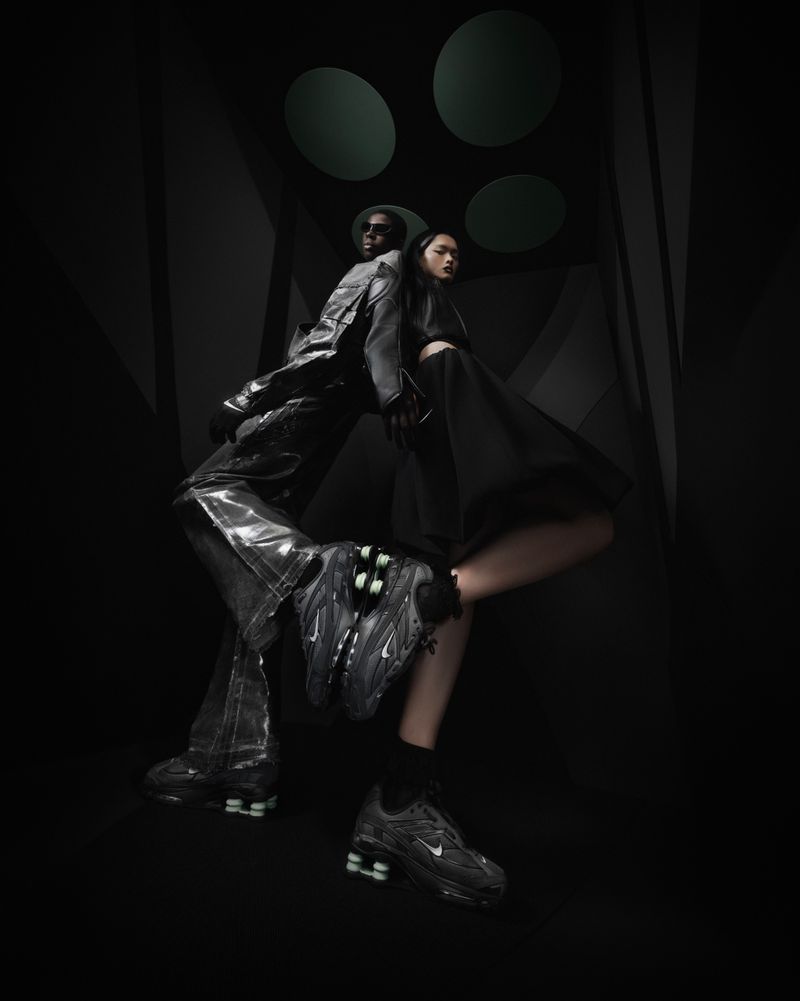 Editorial fashion shot of a Black adult male and an Asian adult female posing with footwear in a dark studio setting.