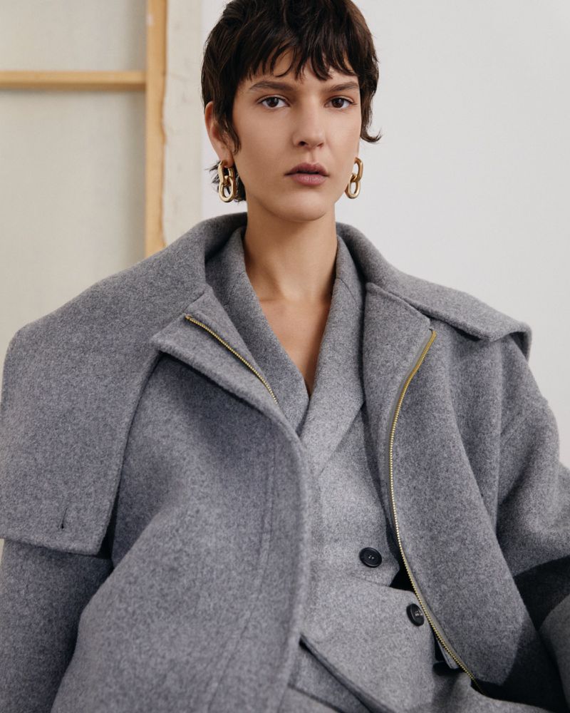A woman poses indoors wearing a grey wool coat and matching suit with gold hoop earrings, against a neutral background with natural light.