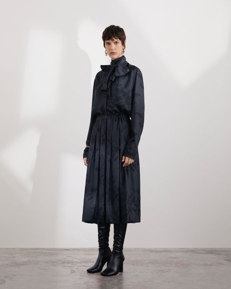 Model wearing a dark patterned long-sleeve blouse with a bow and matching pleated midi skirt against a plain studio background.