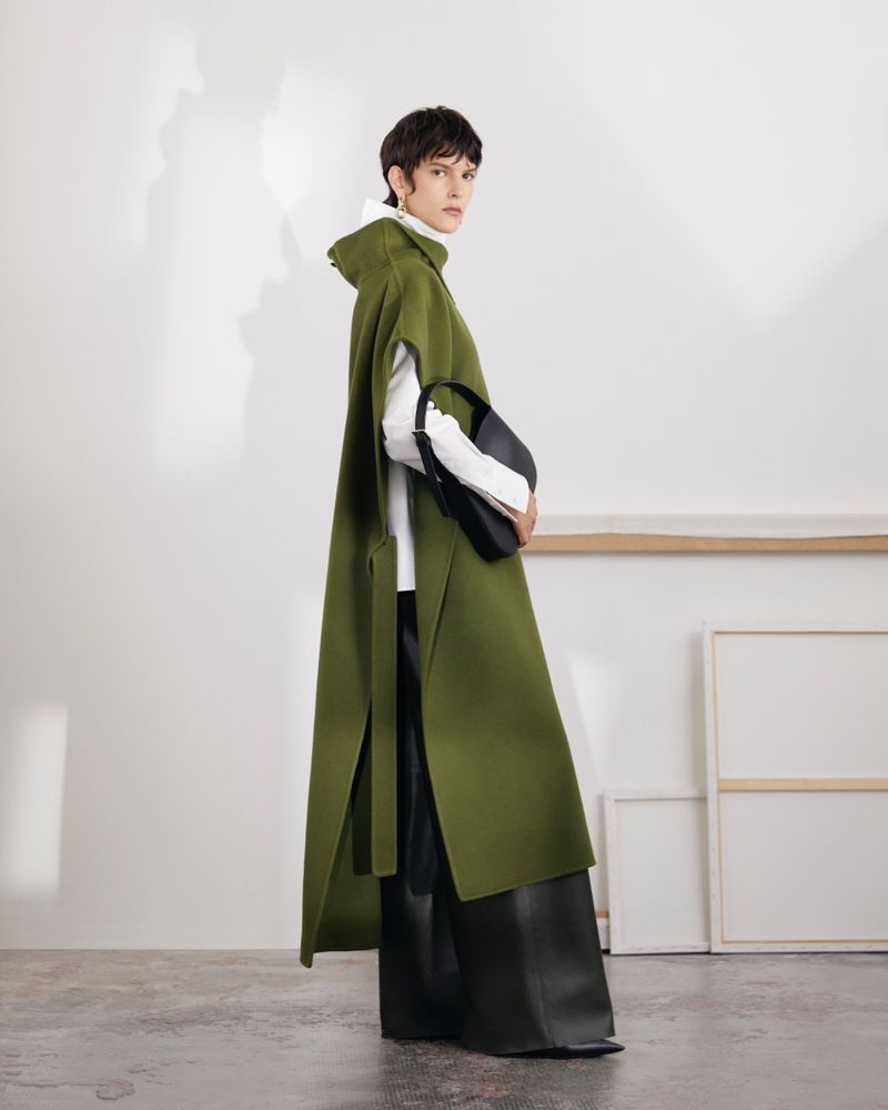A woman in an olive green hooded cape holds a black purse in a minimalist studio setting, showcasing contemporary fashion.