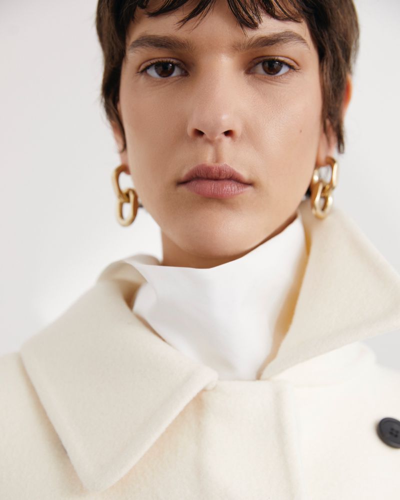 Close-up portrait of an adult woman with short brunette hair, wearing gold earrings, white top, and a light beige wool coat.