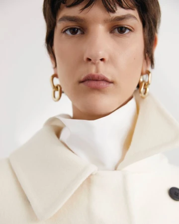 Close-up portrait of an adult woman with short brunette hair, wearing gold earrings, white top, and a light beige wool coat.