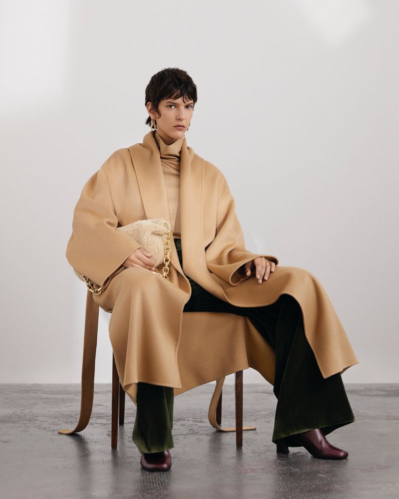 A woman with short brown hair sits in a studio on a wooden chair wearing a tan coat, dark pants, and holding a chain purse.