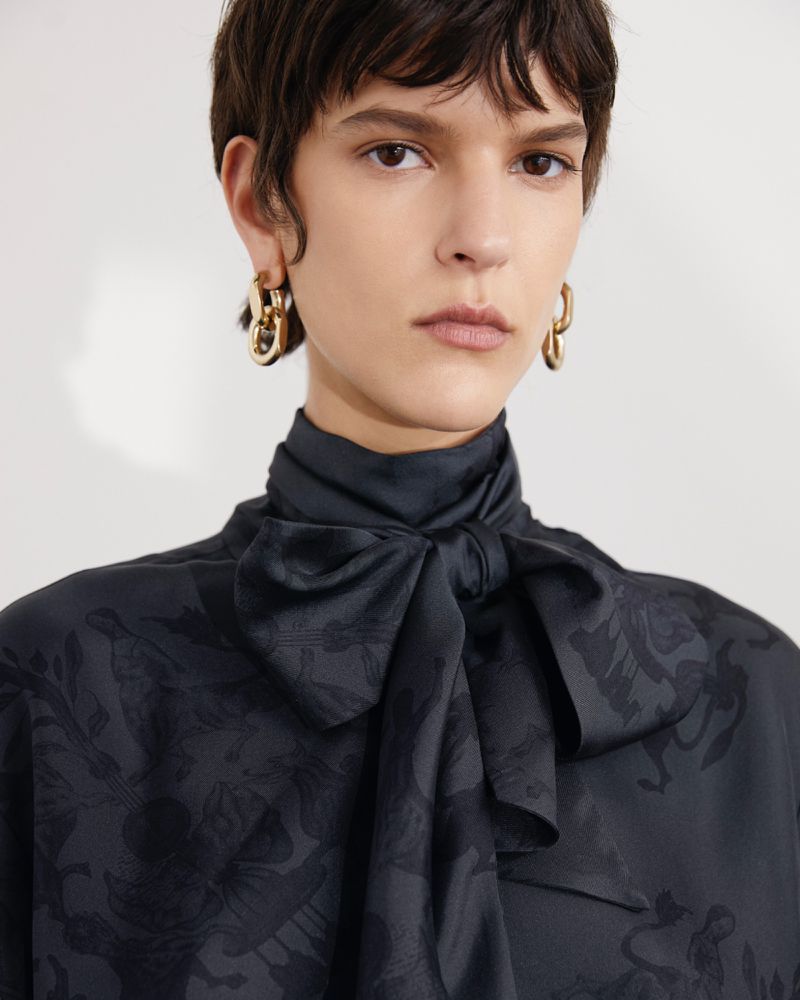 Close-up of a woman with short brown hair wearing gold earrings and a dark patterned blouse with a bow at the neck. Fashion shot.