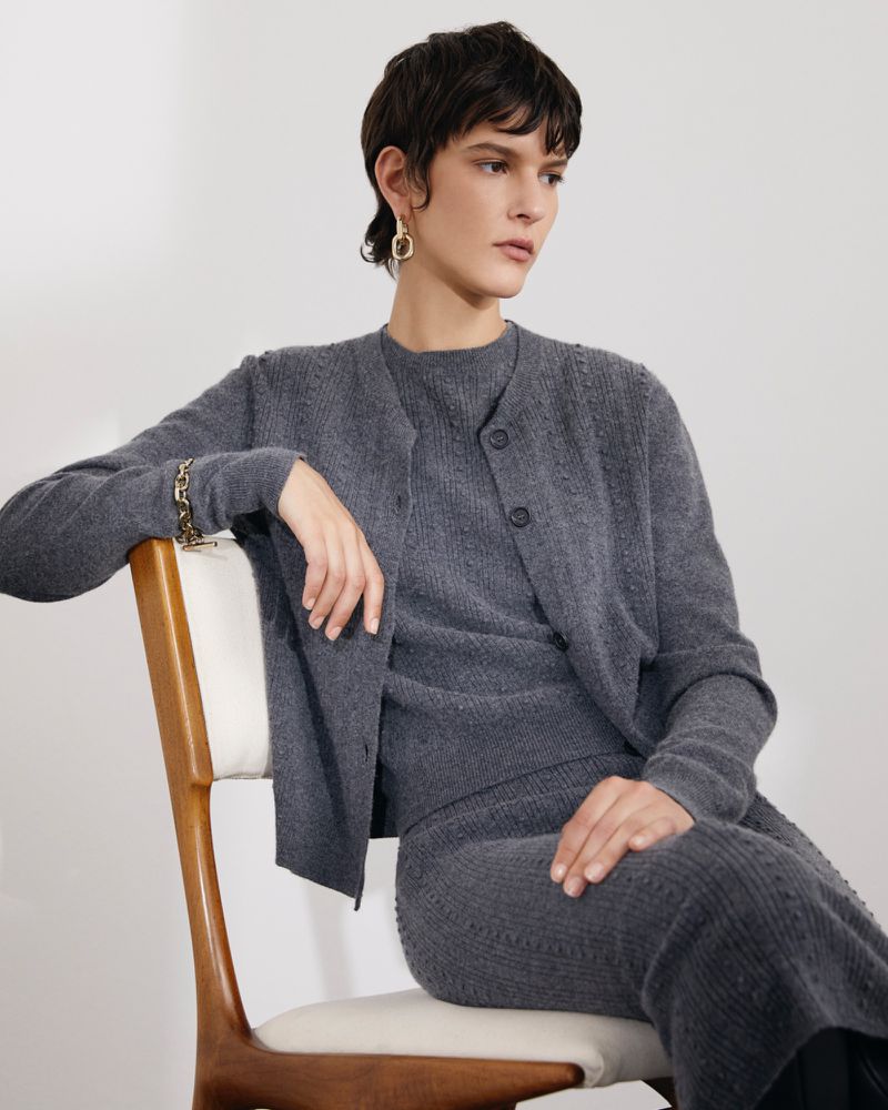 Adult woman in a gray knit sweater and skirt sitting in a chair in front of a white wall wearing gold earrings and bracelet.