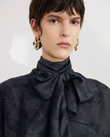 Close-up portrait of a woman wearing a black patterned silk blouse with a large bow collar and gold link earrings against a light background