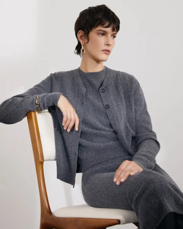 Model with short dark hair wearing a matching grey knit cardigan and skirt seated on a wooden chair against a plain white wall