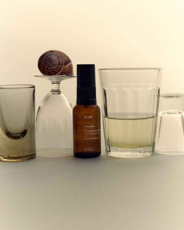 Still life featuring a bottle of Klur Immersion serum, a snail shell on top of a wine glass, and an assortment of glassware.