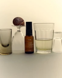 Klur skincare serum bottle arranged in a still life with glassware and a snail shell on a neutral surface.
