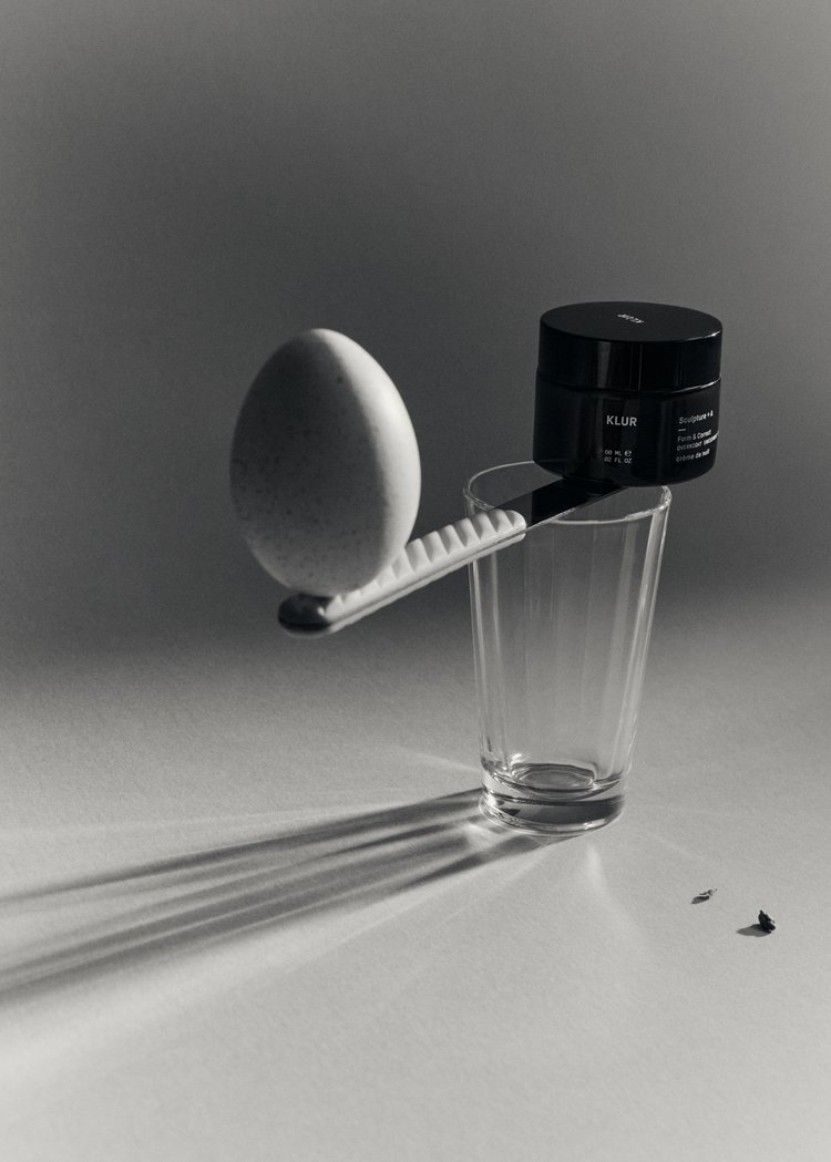Still life shot featuring an egg, a black cosmetic container, and a drinking glass arranged on a white surface with distinct shadows.