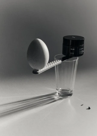 Still life shot featuring an egg, a black cosmetic container, and a drinking glass arranged on a white surface with distinct shadows.