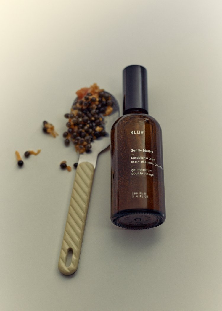 Brown glass bottle of Klur Gentle Matter cleanser, with papaya seeds on a spoon against a plain background in a studio shot.