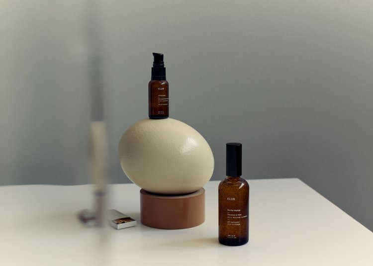Two brown glass bottles are arranged on a white table with a cream-colored decorative sphere in the background against a neutral wall.