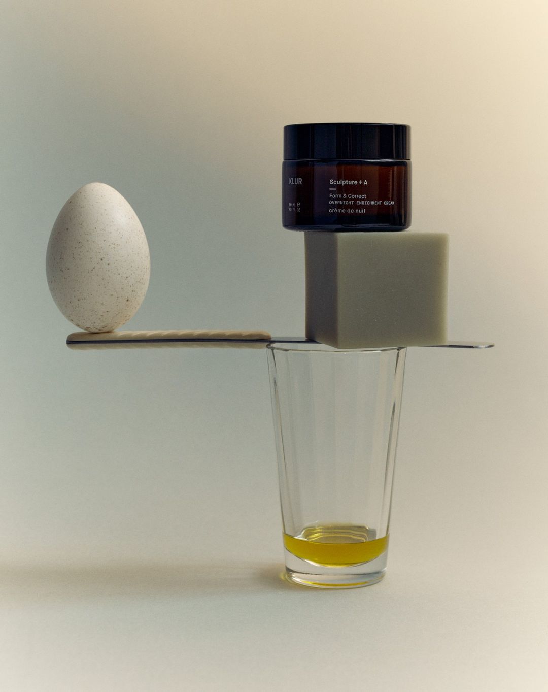 Elevated eye-level still life of balancing beauty product container, bar of soap, egg, eating utensil, and drinking glass with yellow liquid.