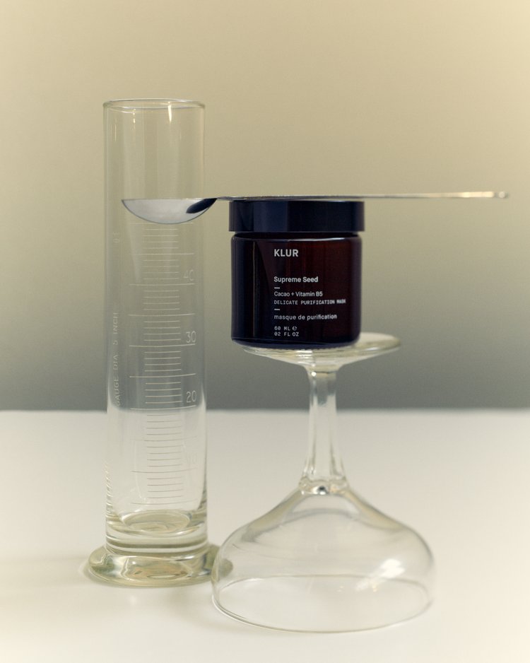 KLUR Supreme Seed mask in a dark glass jar balancing on an upside-down clear glass with a spoon and graduated cylinder.