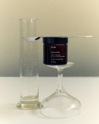 Klur skincare jar resting on an inverted wine glass beside a tall graduated glass cylinder on a white surface.