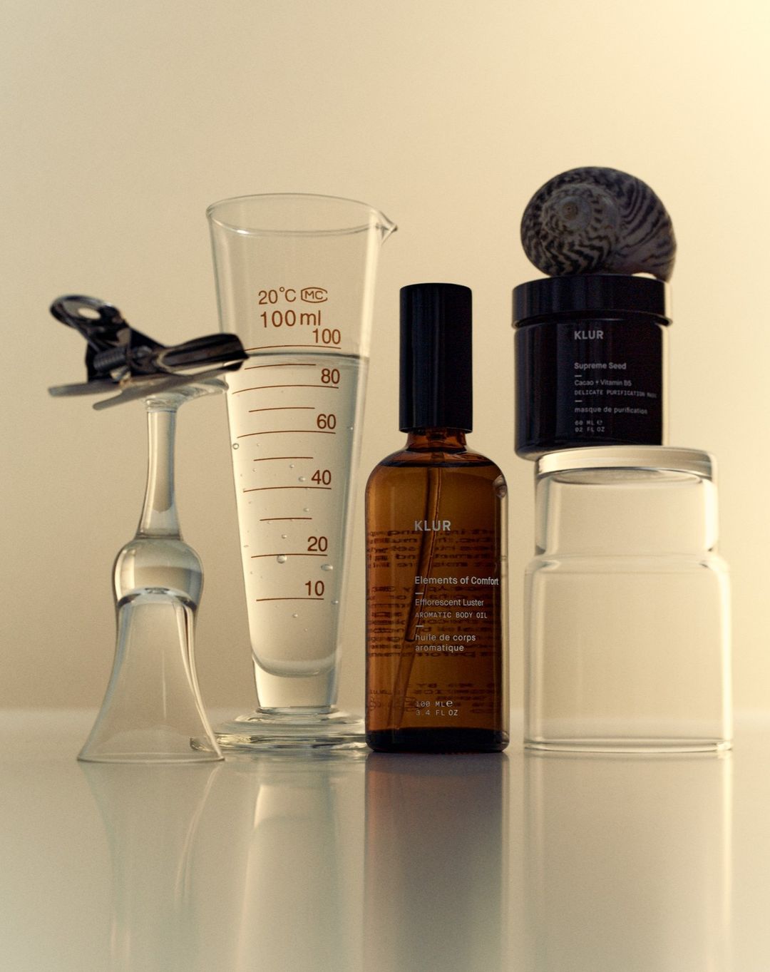 Still life shot of Klur skin care products, clear glass measuring cup with water, glass jars, clam shell on white backdrop.
