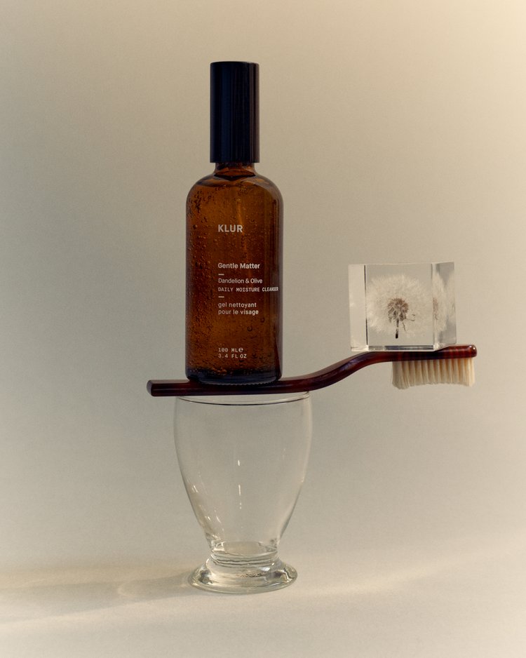 Aesthetic still life of Klur Gentle Matter cleanser bottle, hairbrush, and cube with dandelion on an off-white background.