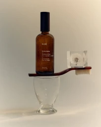 Amber glass skincare bottle and a toothbrush balancing on a clear glass goblet against a neutral wall background