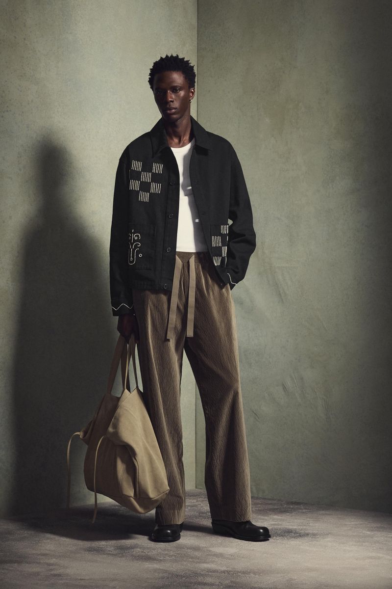 Adult male in black jacket and tan pants holding beige tote bag in studio with neutral backdrop, fashion editorial style.