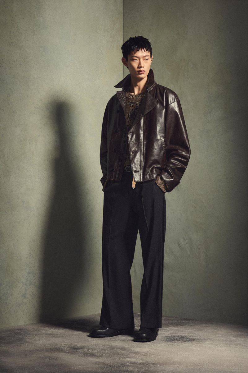 Asian male model in dark leather jacket and black wide-leg trousers poses in a photo studio with a grunge-textured wall backdrop.