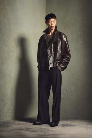 Asian male model in dark leather jacket and black wide-leg trousers poses in a photo studio with a grunge-textured wall backdrop.
