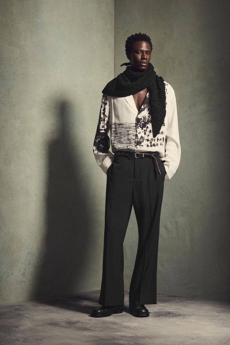 Dark-skinned male model in off-white patterned shirt, black pants and scarf poses indoors against textured green wall with shadow.