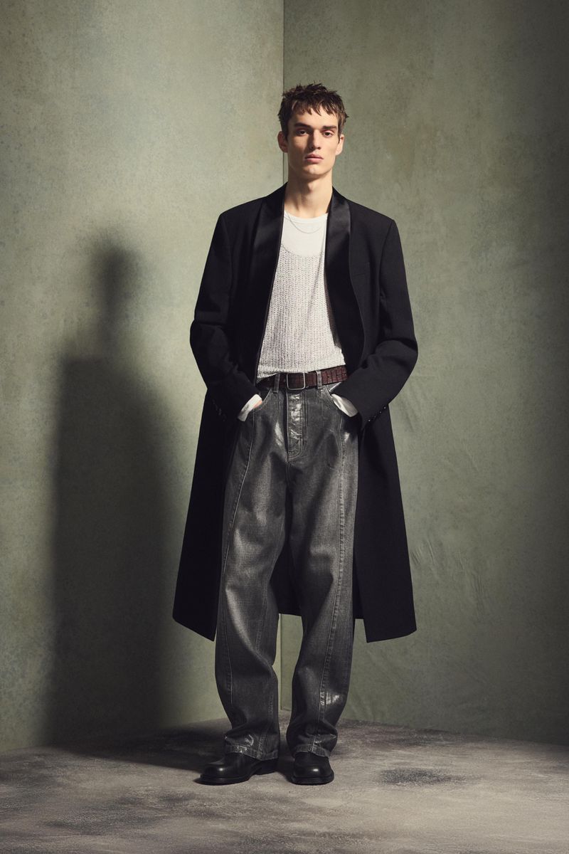 Fashionable man in a black overcoat and grey denim trousers poses in front of a green wall in a full-length studio portrait.