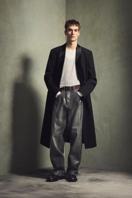 Fashionable man in a black overcoat and grey denim trousers poses in front of a green wall in a full-length studio portrait.